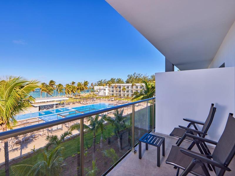 Balkon mit drei Stühlen und Blick auf Pool, Palmen und Hotelgebäude bei klarem Himmel.