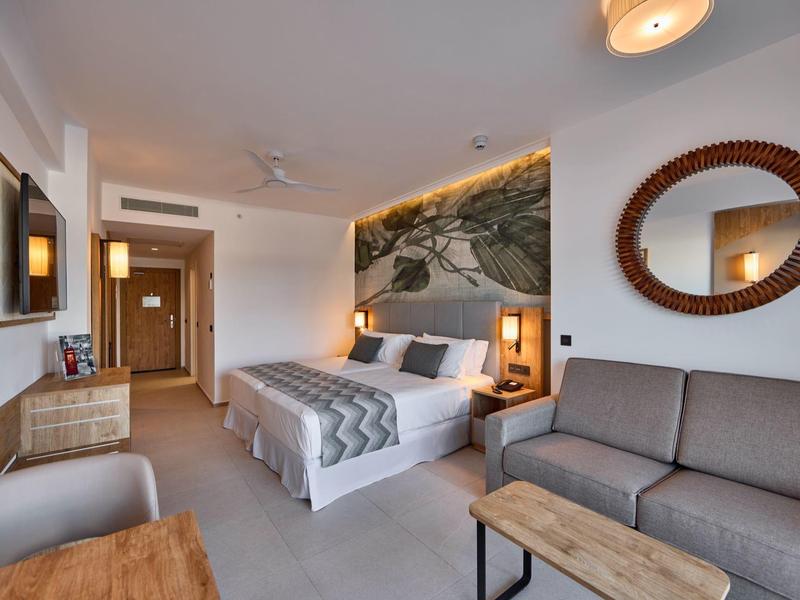 Helles Hotelzimmer mit Doppelbett, grauem Sofa, rundem Spiegel und Landschaftsbild an der Wand.