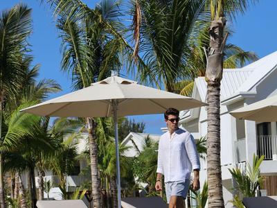 Ein Mann in weißer Freizeitkleidung am Pool neben Palmen und Liegestühlen unter Sonnenschirmen.