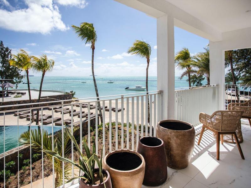 Balkon mit Stühlen und Töpfen mit Blick auf das Meer und Palmen bei klarem Himmel.