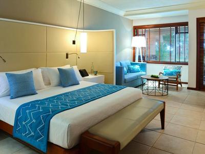 Modern hotel room with large bed, blue pillows, and comfortable armchairs by windows.