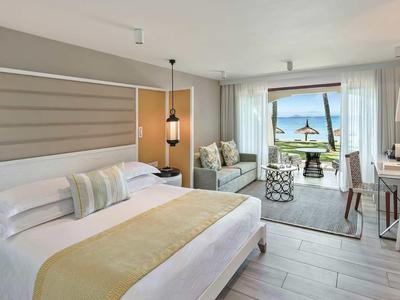Bright hotel room with large bed, seating area, and sea view through balcony door.