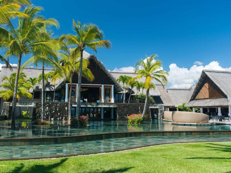 Luxury bungalows with thatched roofs and palm trees by a clear pool under a blue sky.