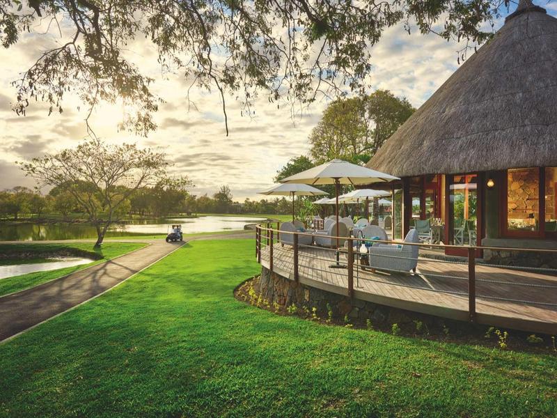 Round hotel with thatched roof and terrace next to green lawn and lake at sunset.