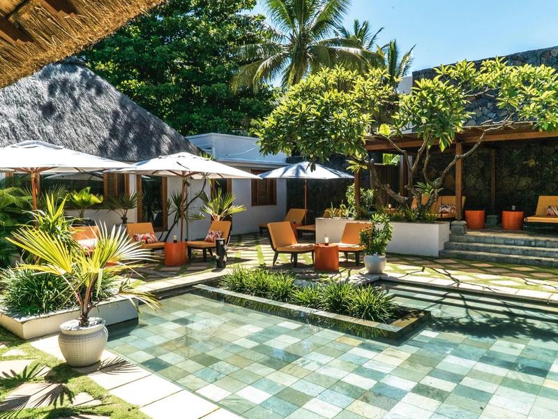 Hotel entrance with pools, chairs, umbrellas surrounded by plants and trees.