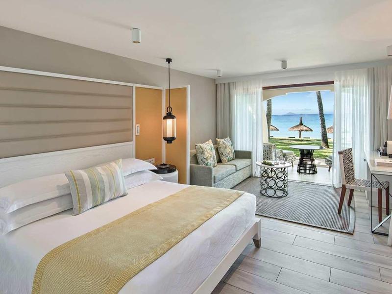 Bright hotel room with large bed, seating area, and sea view through balcony door.
