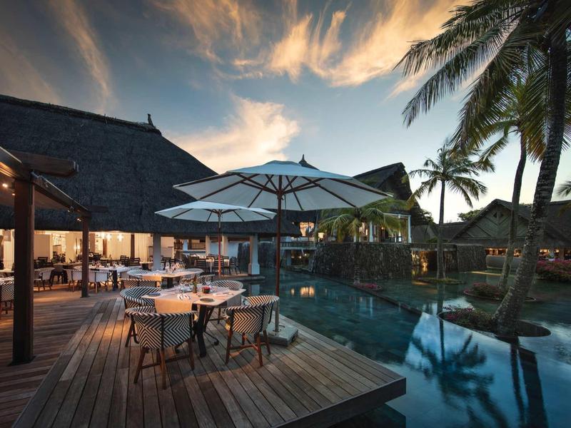 Evening poolside bar at resort with tables, umbrellas, and palm trees.