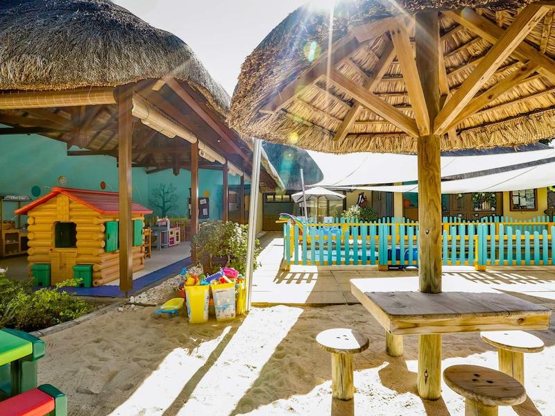 Children's area with sandbox, wooden furniture, and playhouse under thatched roofs outdoors.