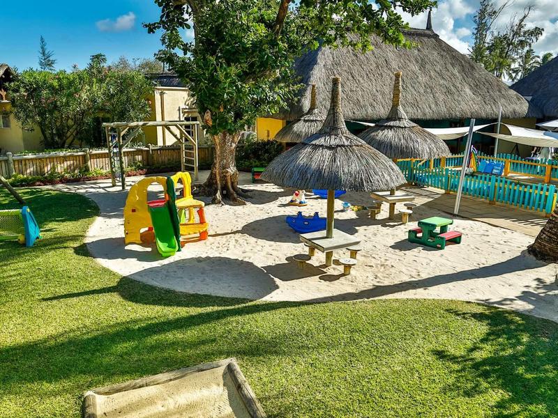 Children's playground with sand area, swings, and straw sunshades in a tropical garden.