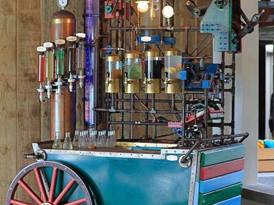 Barevný kovový robot s koly stojí na dřevěné podlaze vedle dřevěné stěny.