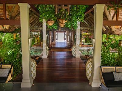 Vstupní prostor tropického resortu s dřevěnou podlahou, bílými sloupy a zelenými rostlinami.