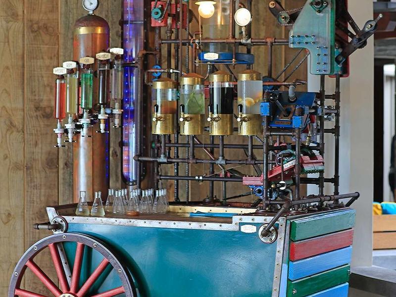 Barevný kovový robot s koly stojí na dřevěné podlaze vedle dřevěné stěny.