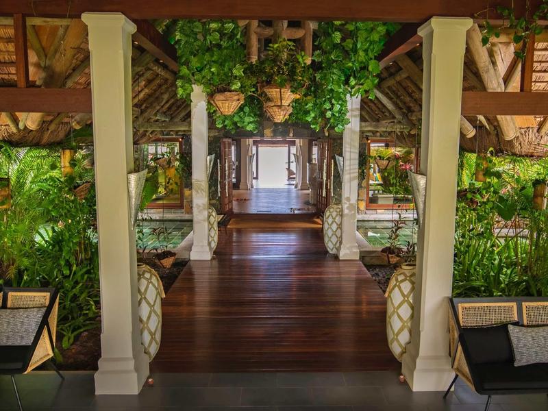 Vstupní prostor tropického resortu s dřevěnou podlahou, bílými sloupy a zelenými rostlinami.