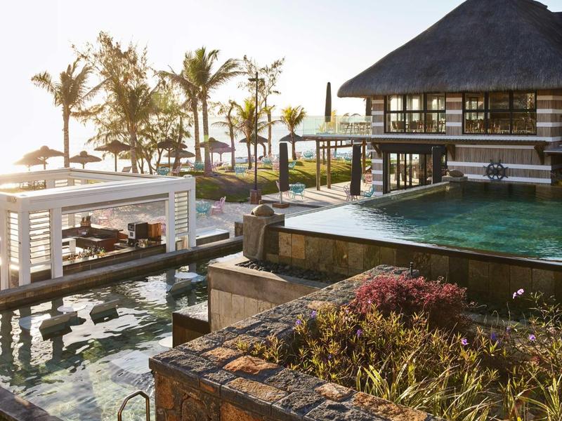 Luxusní hotel s infinity bazénem, pavilonem a palmami u moře při západu slunce.