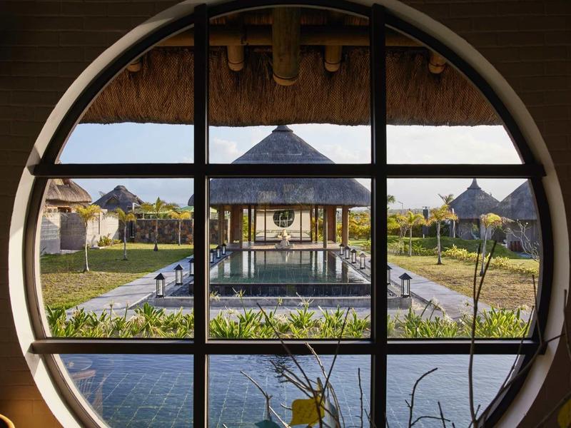 Pohled přes kulaté okno na zahradu s pavilonem a cestami v tropickém hotelu.