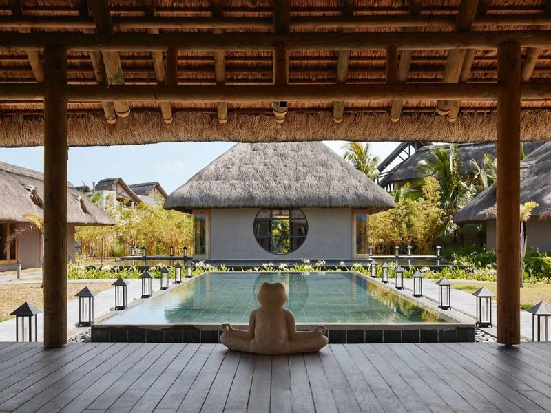 Výhled z krytého prostoru na bazén obklopený bungalovy a tropickou vegetací.