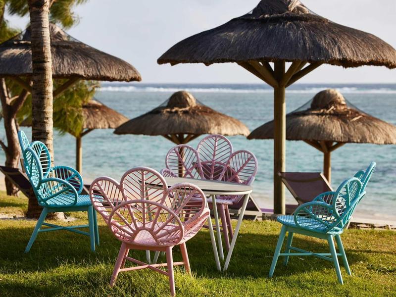 Barevné židle a stoly pod slaměnými slunečníky na pláži s výhledem na moře.