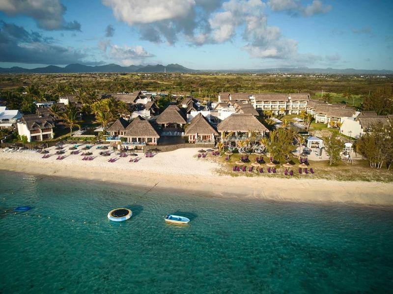 Přímořské letovisko s bungalovy se střešní krytinou z doškových střech, slunečníky a loděmi v průzračné modré vodě.