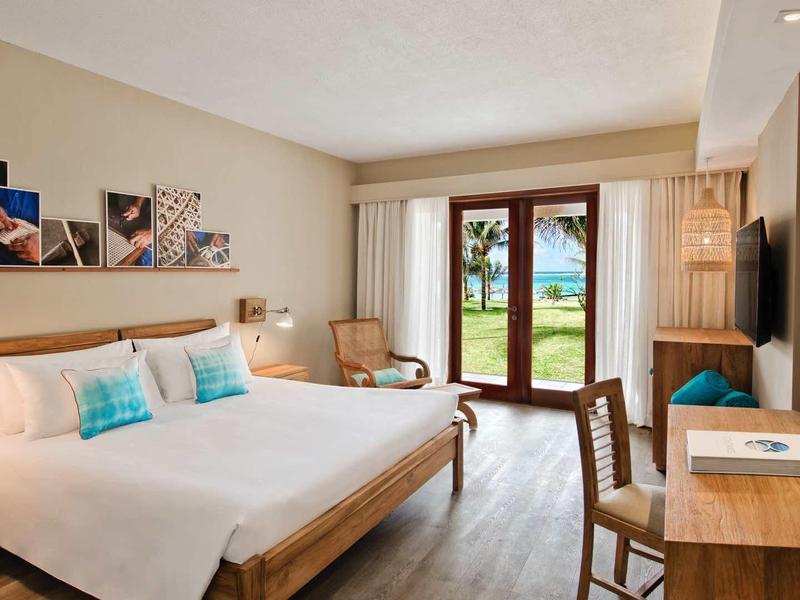 Světý hotelový pokoj se dvěma postelemi, psacím stolem a výhledem na zahradu přes velké balkonové dveře.