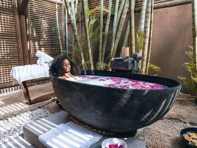 Woman relaxing in large black outdoor bathtub surrounded by plants and wooden walls.