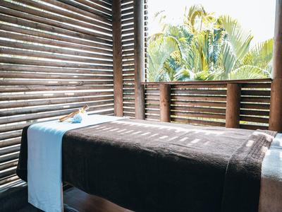 A spa massage table with dark cover and light blue towel roll in a room with wooden slats and palm view.