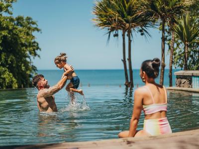 Vater hebt Kind im Pool, Frau sitzt am Beckenrand mit Blick aufs Meer und Palmen.