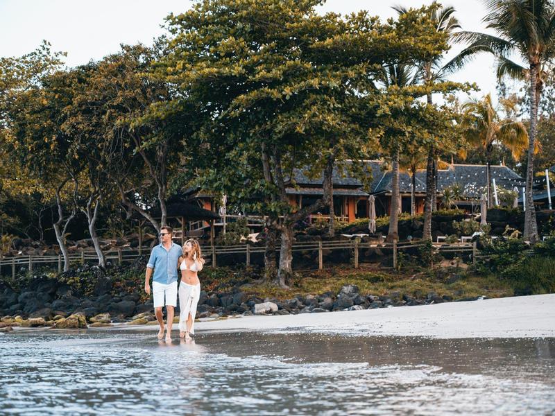 Couple walking on the beach near tropical vegetation and wooden huts.