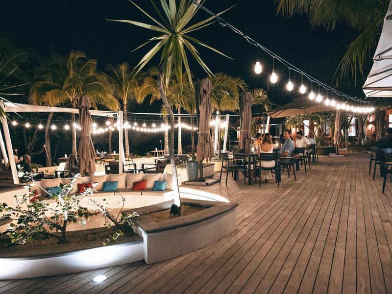 Lively hotel terrace with wooden floor, string lights, and palm trees at night.