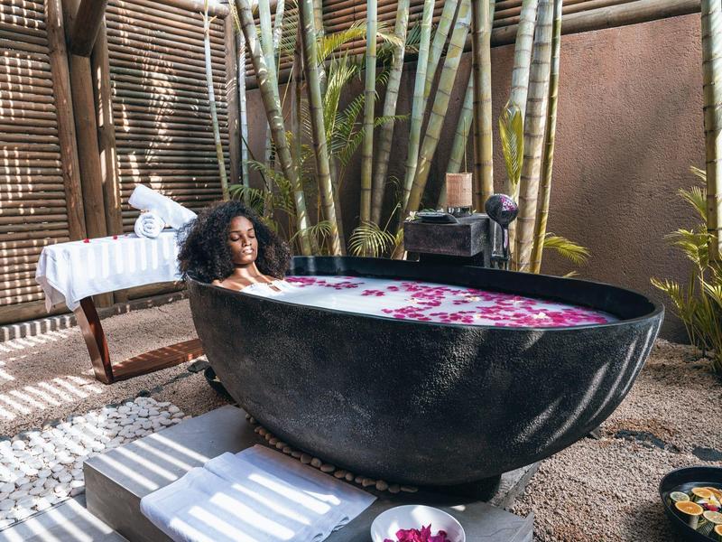 Woman relaxing in large black outdoor bathtub surrounded by plants and wooden walls.