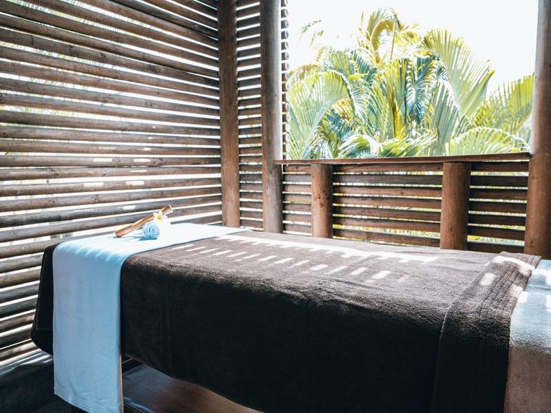 A spa massage table with dark cover and light blue towel roll in a room with wooden slats and palm view.