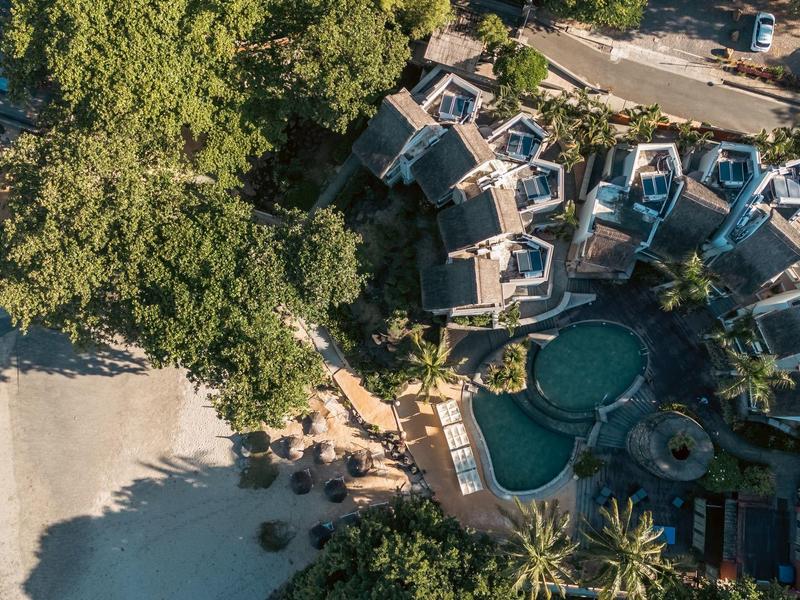 Aerial view of a tropical resort with pool, bungalows, and sandy beach by clear water.