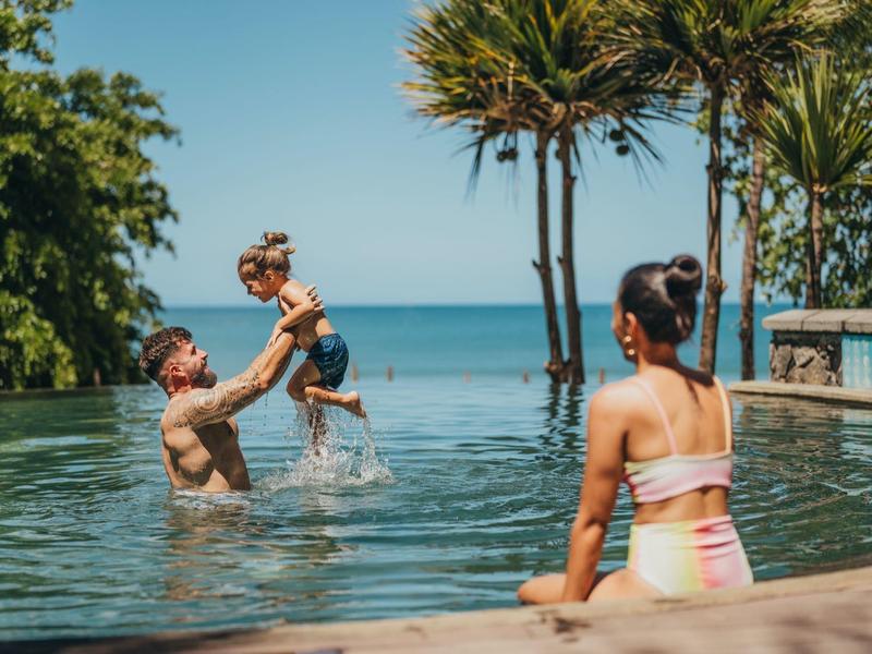 Family relaxing at an infinity pool overlooking the sea with tropical plants.