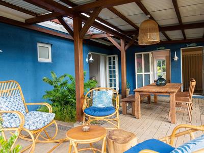 Gemütliche Terrasse mit blau gestrichener Wand, Holztisch, Stühlen und Korbmöbeln unter Holzüberdachung.