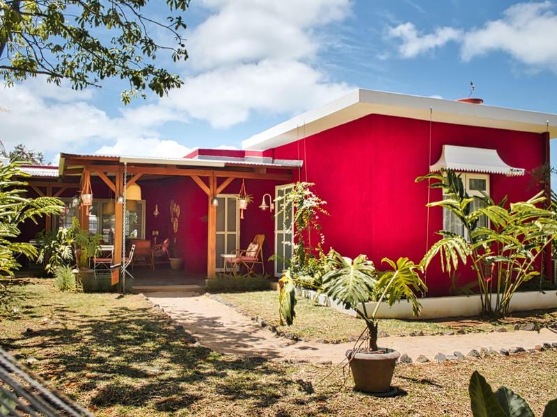 Modernes, hellrotes Haus mit Holzterrasse, Pflanzen im Garten und blauem Himmel im Hintergrund.