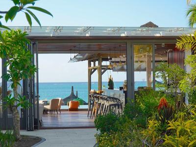 Offener Strandpavillon mit Holzmöbeln, Pflanzen und Blick auf den blauen Ozean bei klarem Himmel.