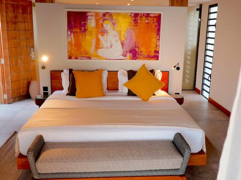 Světlý hotelový pokoj s velkou postelí, barevným obrazem a lavicí u nohou.