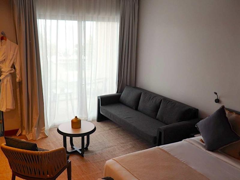 Moderní hotelový pokoj s manželskou postelí, šedou pohovkou, křeslem a kulatým stolem u okna s závěsy.