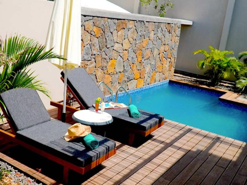 Dvě lehátka s polštáři a nápojem u bazénu s kamennou terasou a rostlinami.