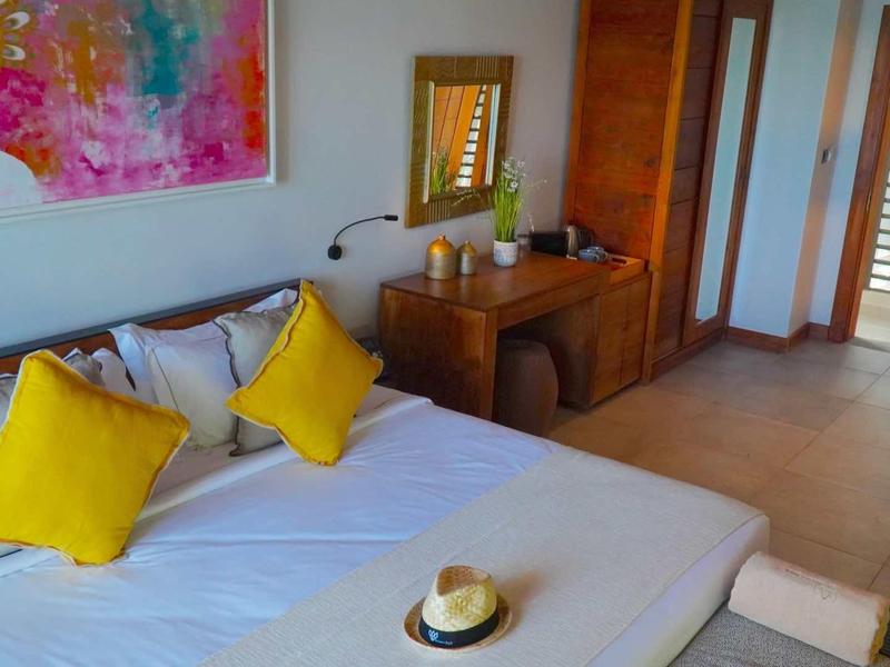 Světlo hotelový pokoj s manželskou postelí, žlutými polštáři a dřevěným nábytkem.