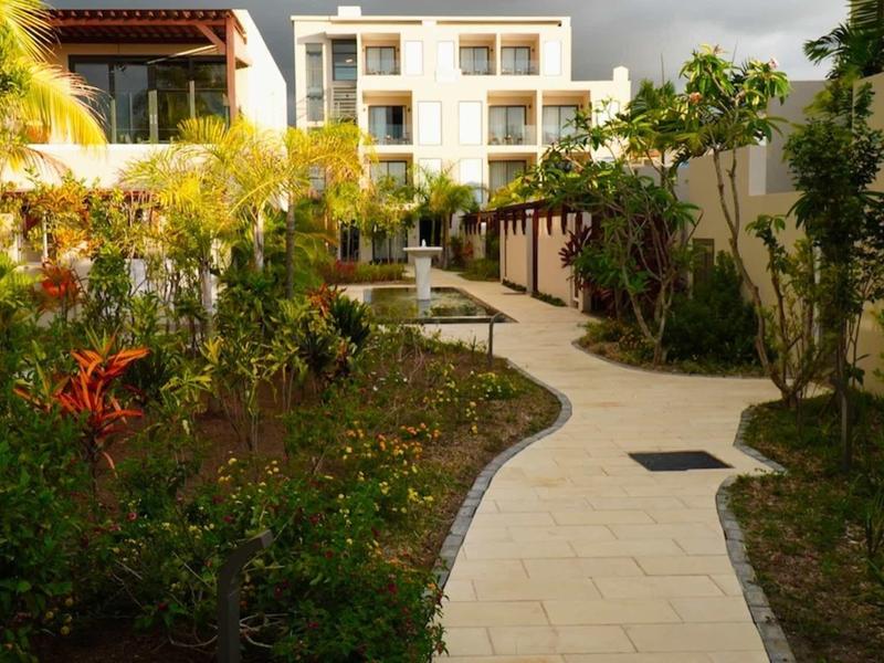 Dlázdená cesta vede zahradou hotelu s rostlinami a stromy pod zataženou oblohou.