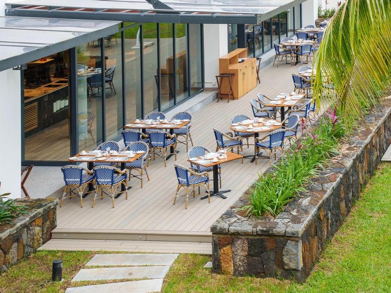 Terasa hotelu s modrými židlemi a stoly vedle skleněné budovy a tropických rostlin.