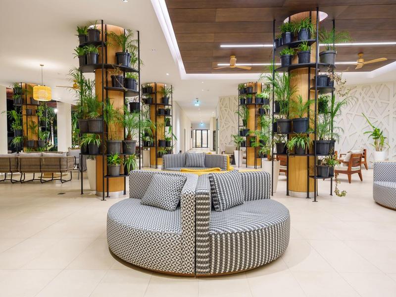 Světlá hotelová hala s kulatým šedým sedacím pufem a zelenými rostlinami na sloupech.