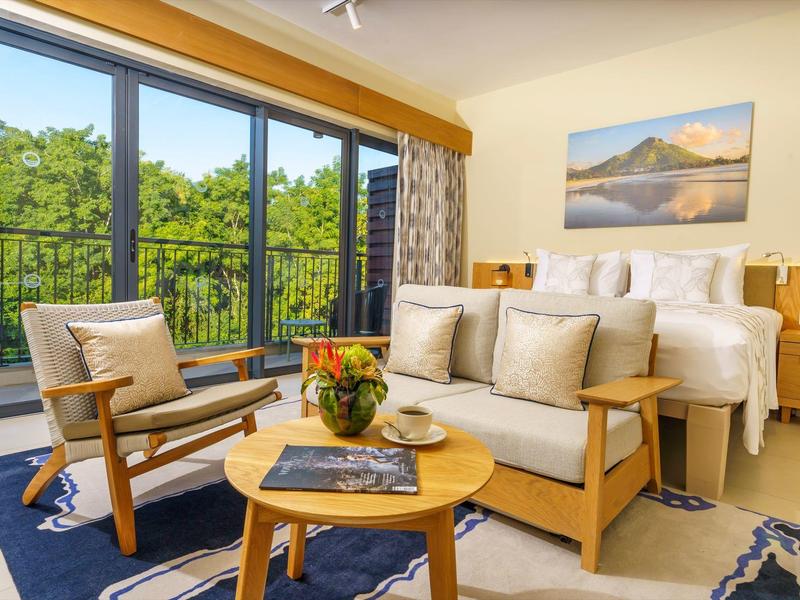 Světý hotelový pokoj s posezením, postelí a výhledem na zelené stromy přes velká okna.