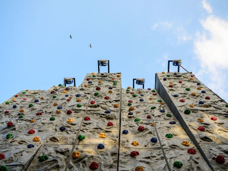 Pohled vzhůru na lezeckou stěnu s barevnými úchyty pod modrou oblohou.