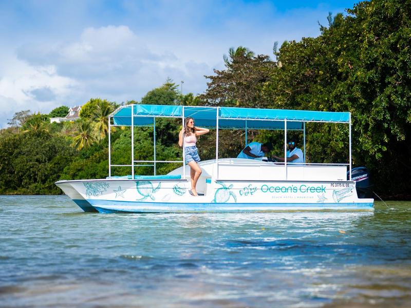 Žena stojí na bílém člunu s modrou střechou na klidné vodě u zeleného břehu.
