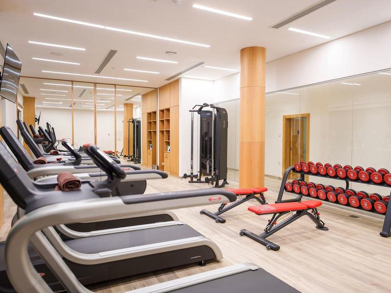 Světle osvětlená posilovna s běžeckými pásy, činkami a lavicí na cvičení.