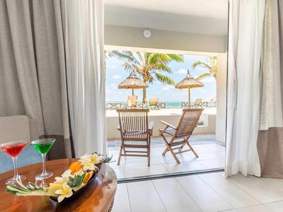 Pohled z hotelového pokoje na terasu se židlemi, stolem a slunečníky u moře.