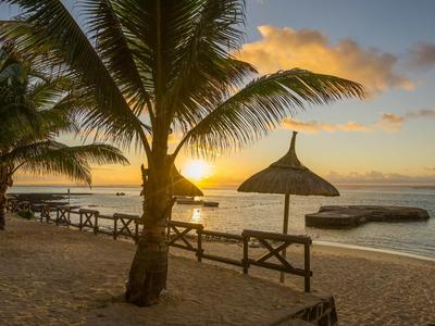 Sonnenuntergang am Strand mit Palmen, Strohschirmen und Sitzgelegenheiten am Wasser.