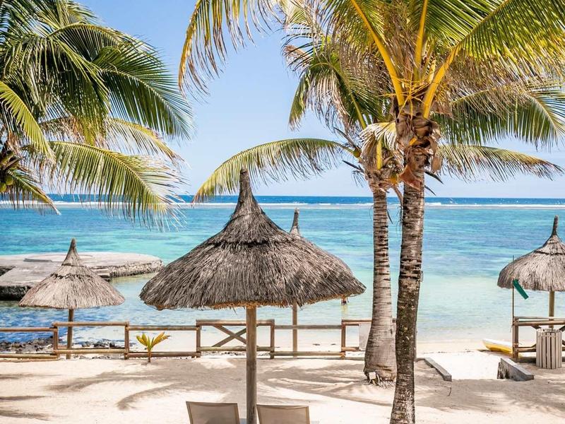 Pláž s palmami, slaměnými slunečníky a lehátky u moře pod modrou oblohou.