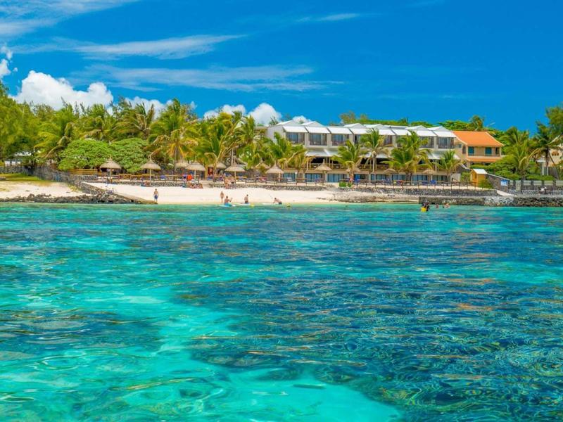 Čirá tyrkysová voda na pláži s palmami a budovami v pozadí.